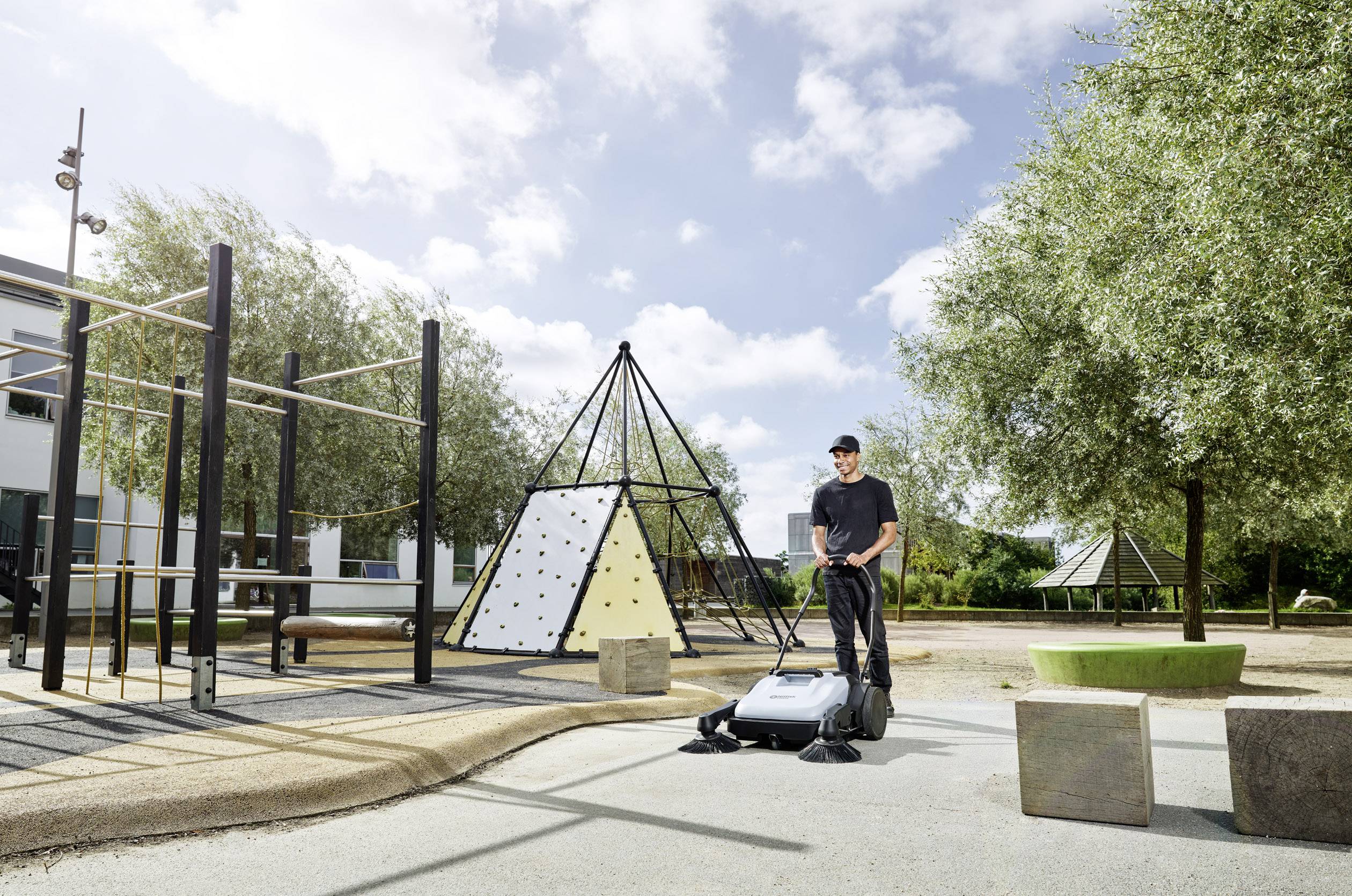 A man is mowing the lawn on a playground next to climbing frames and trees under a blue sky with clouds.