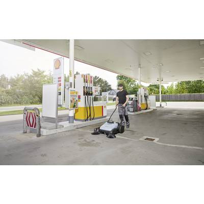 A person is cleaning the floor of a petrol station with a sweeping machine. Fuel pumps and trees are visible in the background.