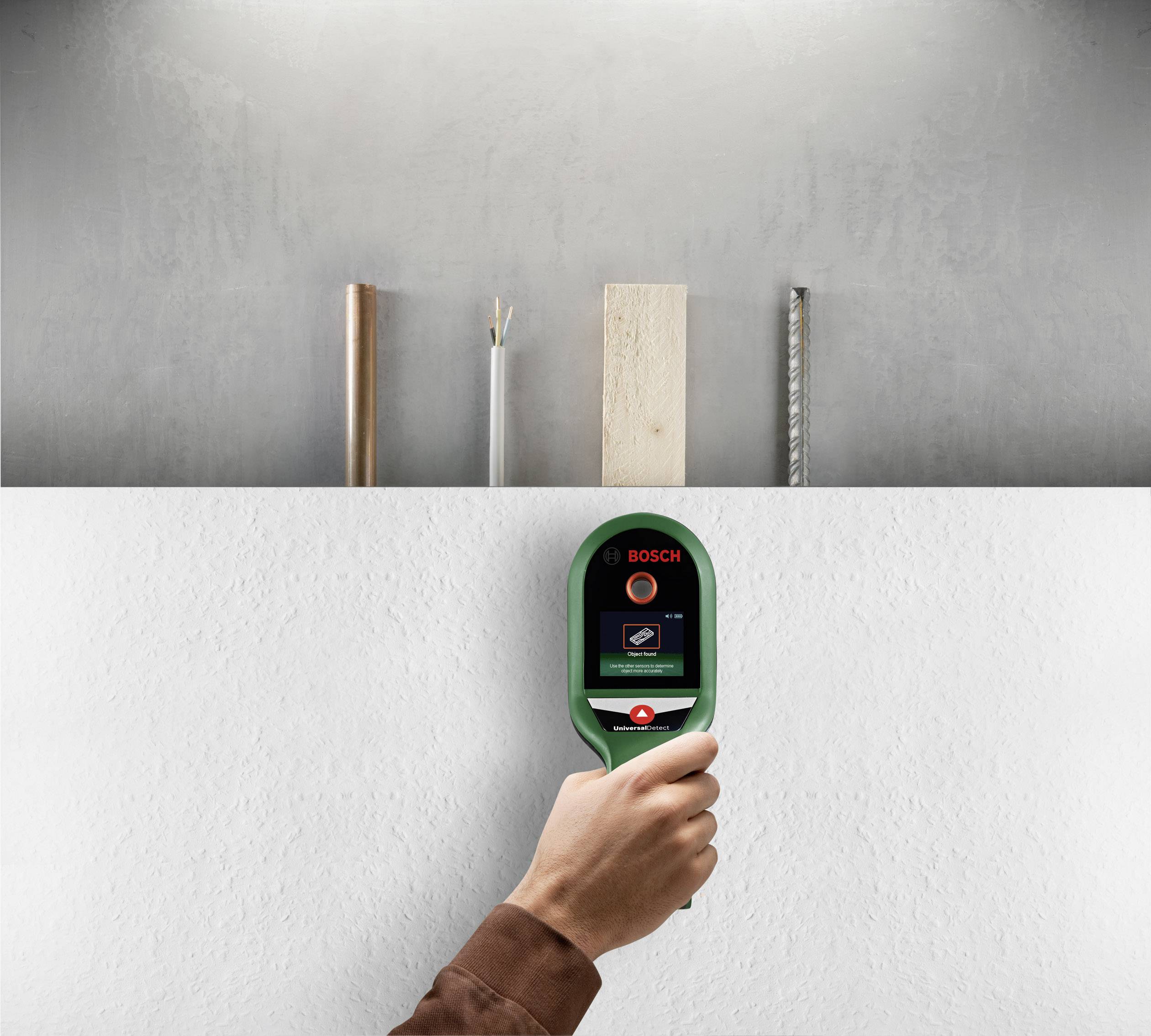 A handheld device for material detection is being used on a wall; metal, wood, and cable materials can be seen in the background.