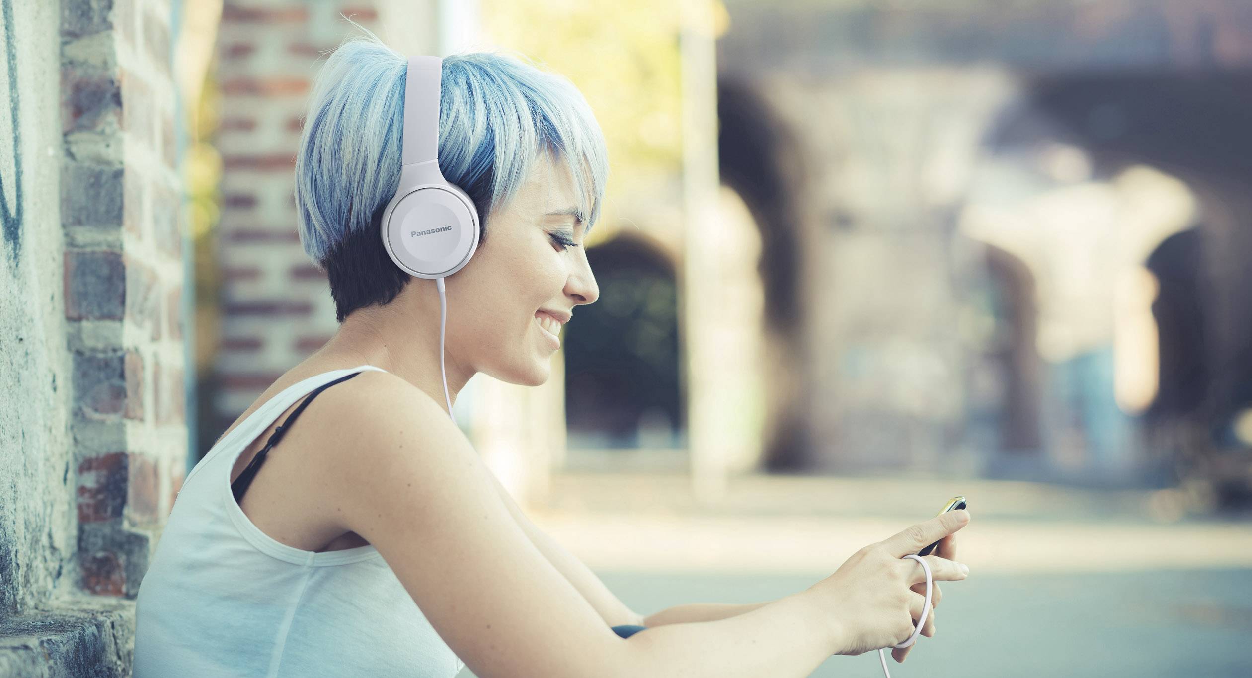 A smiling person with blue hair and headphones is using a smartphone, sitting relaxed outdoors against a blurry city backdrop.