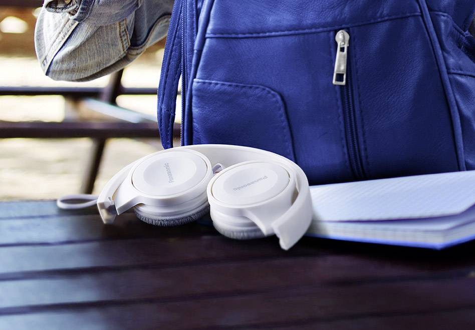 Headphones are lying on a wooden table next to a blue rucksack and a notebook. Blurred benches can be seen in the background.