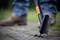 A person wearing blue jeans is removing weeds with a weeding tool from the joints of a paved surface. Blurred green plants are visible in the background.
