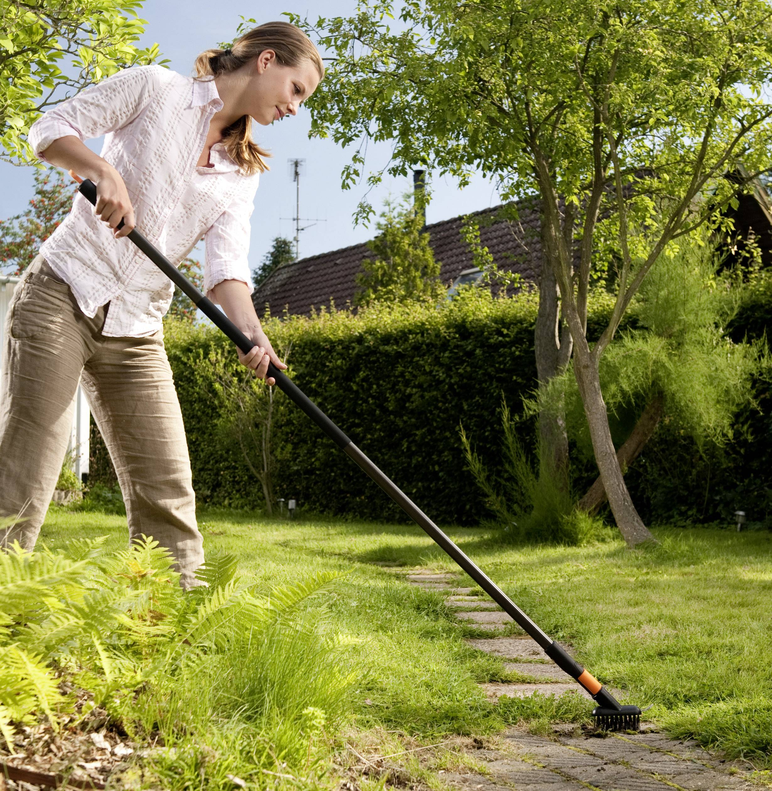 A woman in a garden is using a long-handled tool to remove weeds between paving stones. Trees and bushes are visible in the background.