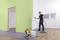 A man is spraying green paint on an interior wall using a spray gun. A paint machine and a bucket are standing on the floor.