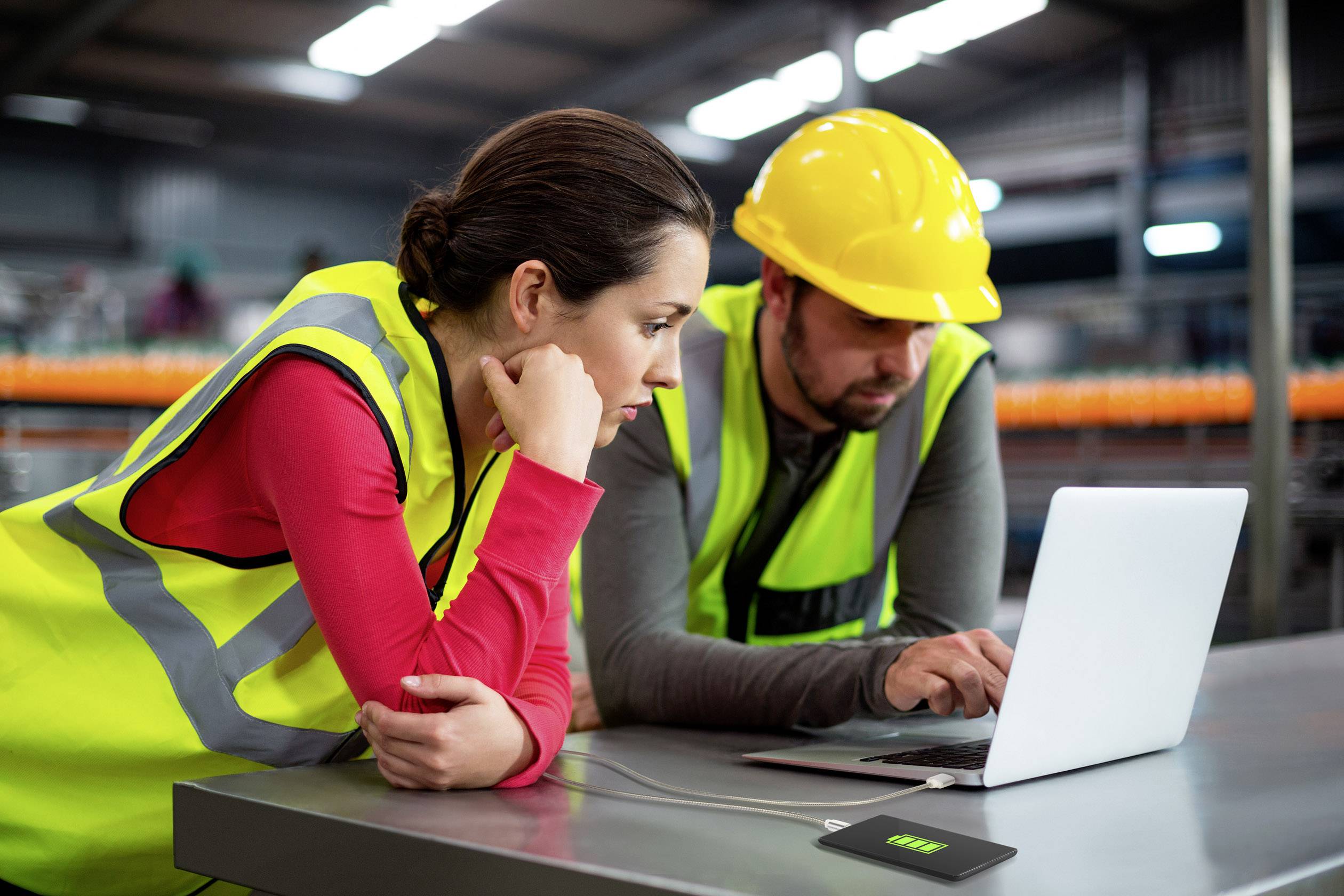 Two individuals in high-visibility vests, a woman and a man wearing a safety helmet, are carefully examining a laptop in a factory setting.