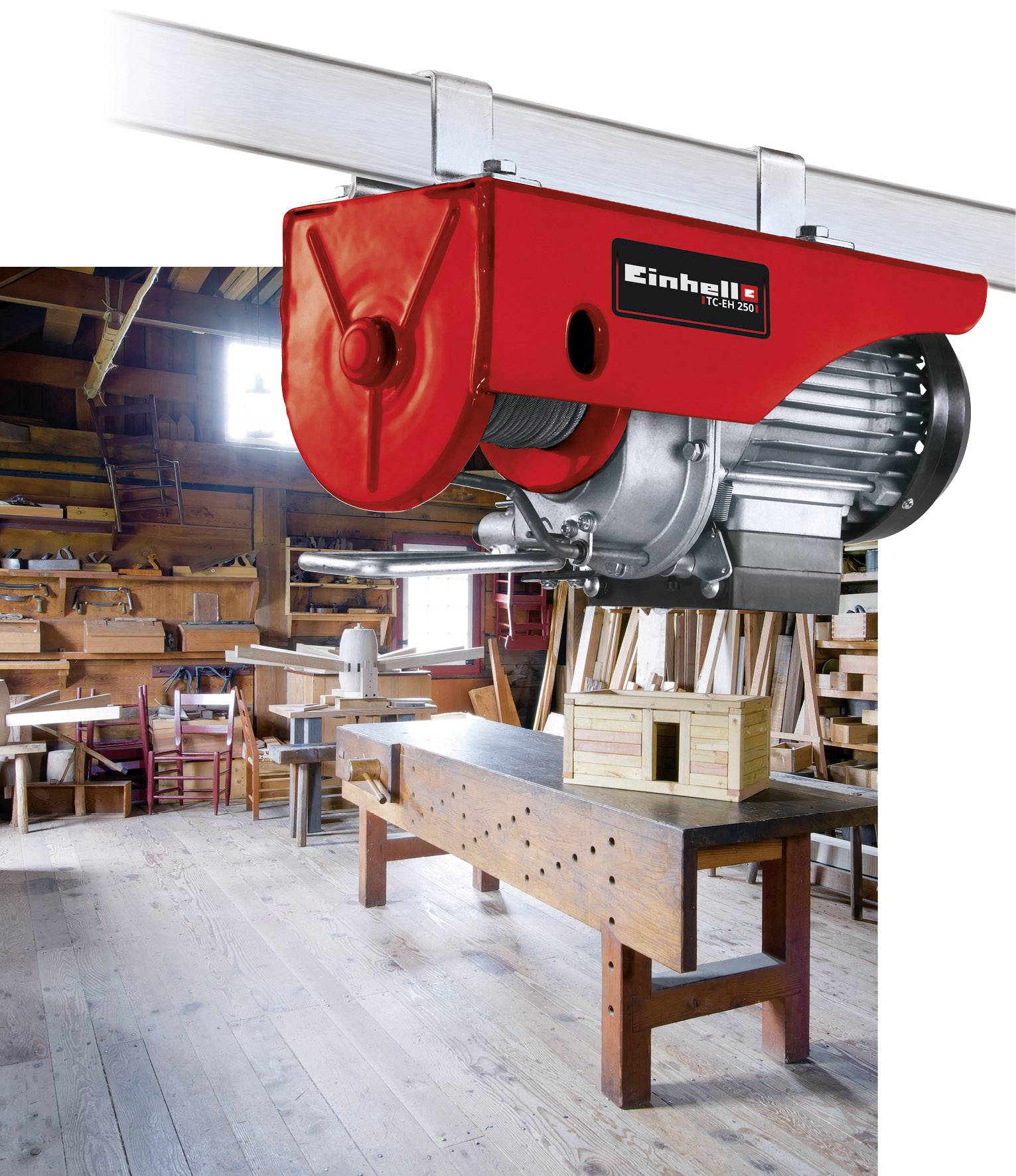 A red winch hangs from a metal pole in a woodworking workshop. Workbenches, wooden shelves, and tools can be seen in the background.