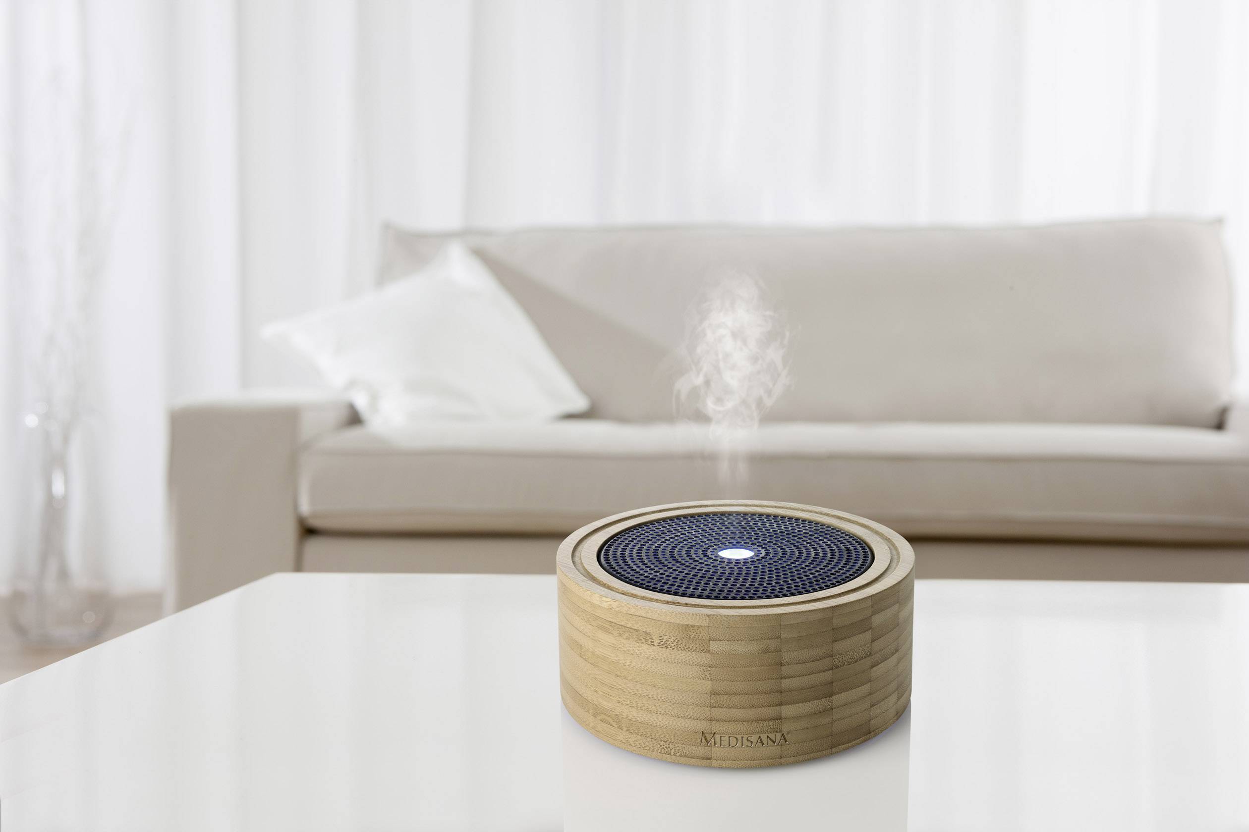 A wooden aroma diffuser sits on a table, releasing vapour. In the background, a sofa can be seen in a modern living room.