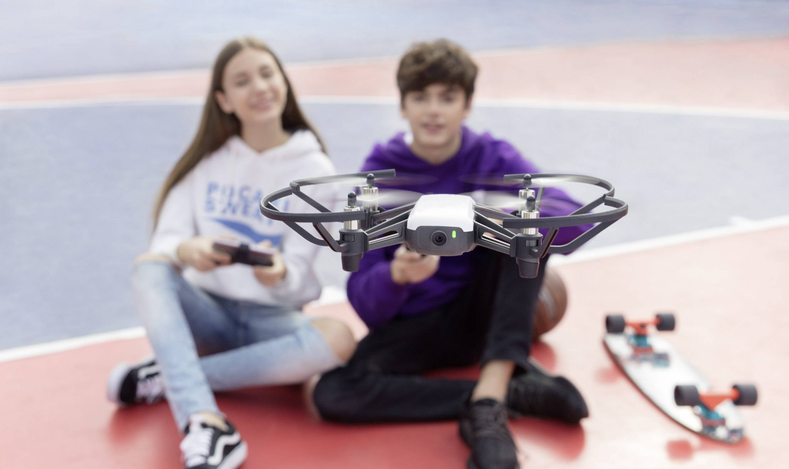 Two teenagers are sitting on a basketball court, controlling a hovering drone. A skateboard can be seen in the background.
