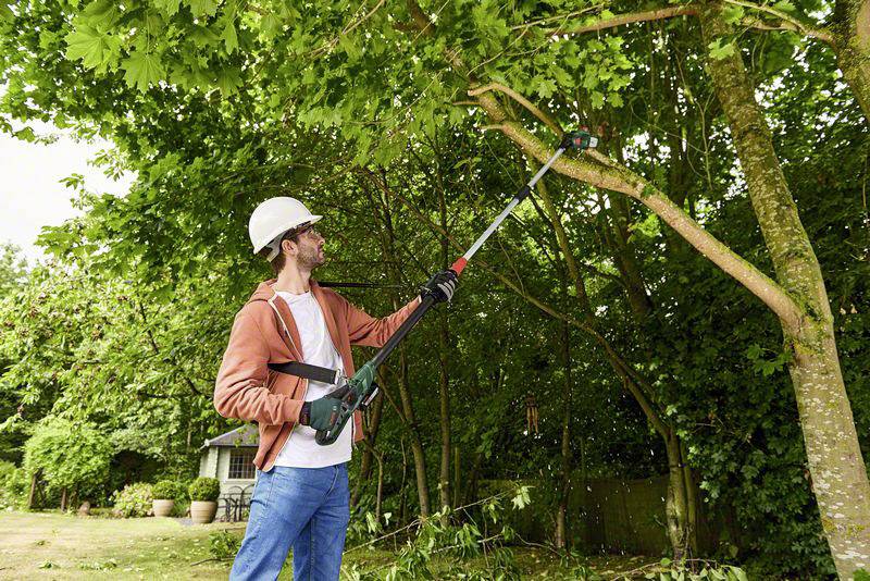 A man wearing safety glasses and a helmet is pruning branches with an electric branch cutter in a garden.