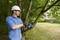 A man in work attire and a safety helmet is using a chainsaw to cut branches from a tree in a garden.