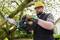 A man in work attire and a hard hat is cutting a branch off a tree using an electric chainsaw.