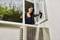 A woman is cleaning an externally mounted air conditioning unit from the inside using a steam cleaner through an open window.
