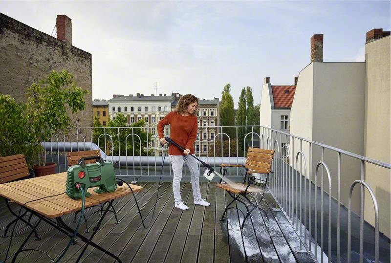 A person is cleaning a wooden floor on a roof terrace with a pressure washer. Urban buildings can be seen in the background.