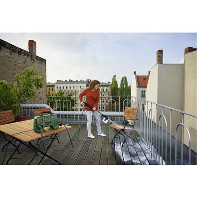 A person is cleaning a wooden floor on a roof terrace with a pressure washer. Urban buildings can be seen in the background.