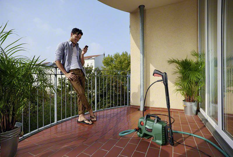 A man is leaning against a balcony, looking at his mobile phone. A pressure washer is standing on the ground, surrounded by plants.