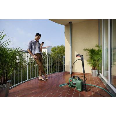 A man is leaning against a balcony, looking at his mobile phone. A pressure washer is standing on the ground, surrounded by plants.
