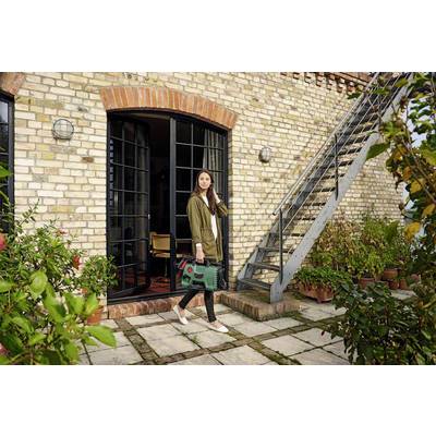 A woman in casual clothing is carrying a green tool bag and standing on a terrace in front of a brick house with a metal staircase.