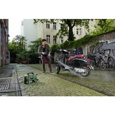 A man is cleaning a motorbike with a pressure washer in a courtyard. Bicycles and graffiti can be seen in the background.