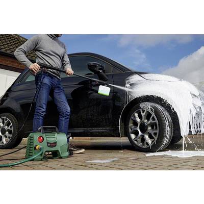 A person is cleaning a car with a pressure washer on a paved courtyard. The car is partially covered with foam.