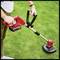 A person is cutting grass in the garden with a red, electric lawn trimmer. The device battery is attached to the trimmer.
