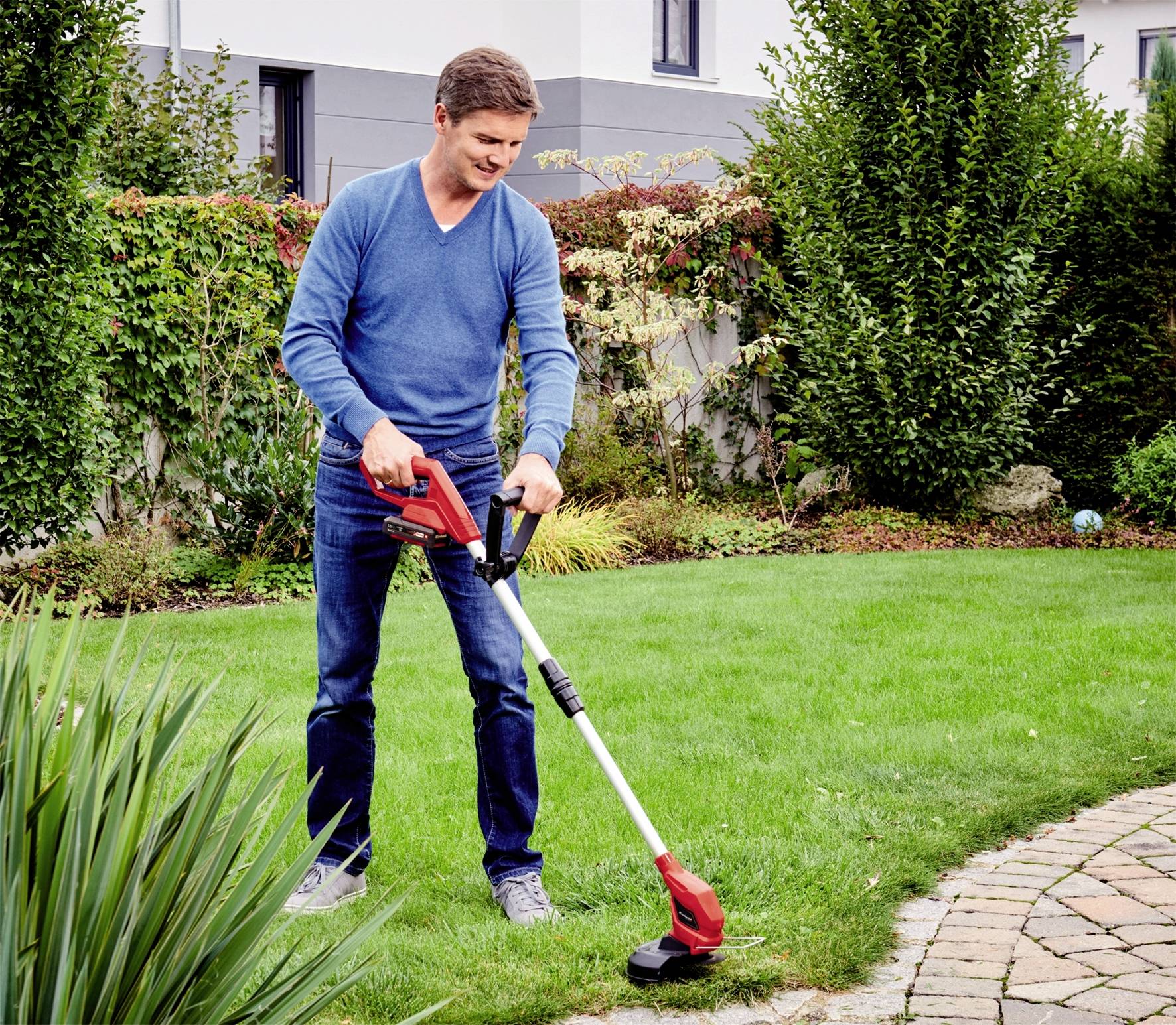 A man is trimming the lawn with an electric lawn trimmer in a garden. Bushes and a house can be seen in the background.