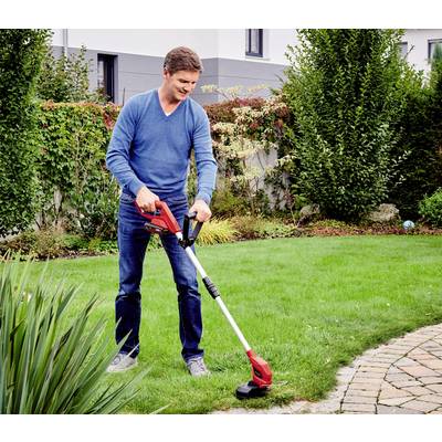 A man is trimming the lawn with an electric lawn trimmer in a garden. Bushes and a house can be seen in the background.