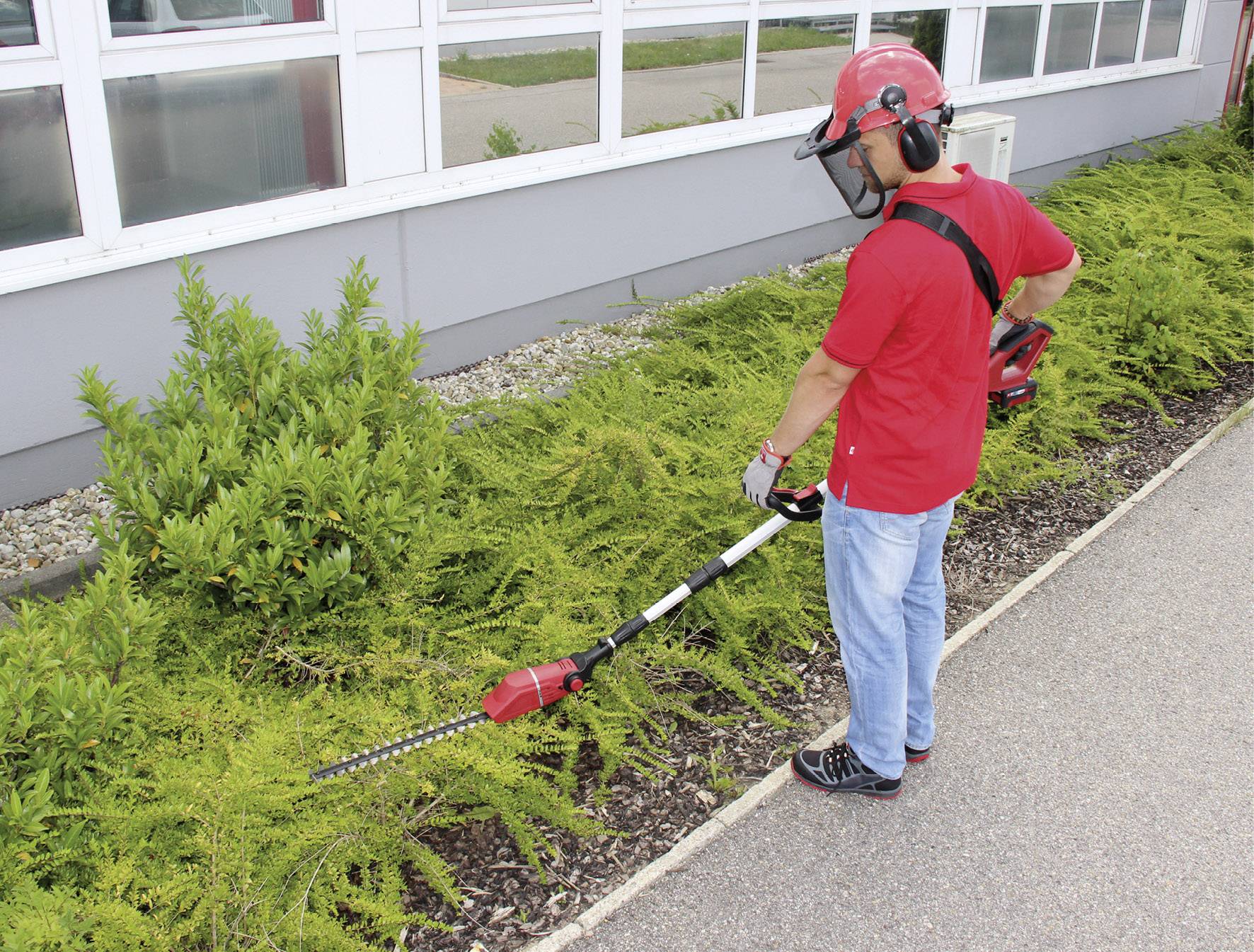 A person in protective clothing is trimming bushes along a building with an electric hedge trimmer.