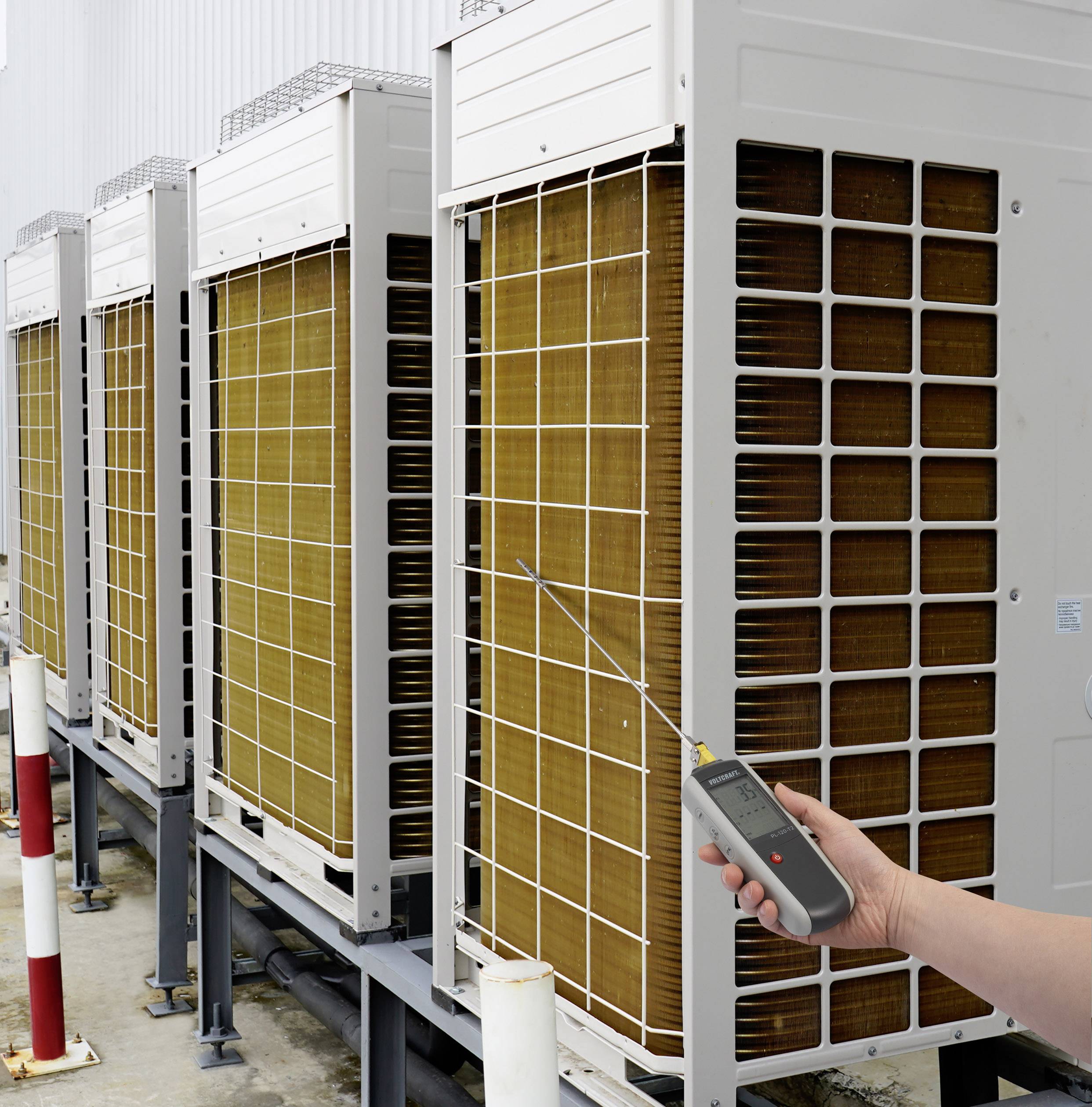 A person is checking air conditioning external units with a digital thermometer. The units are lined up in a row.