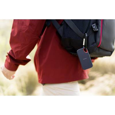 A person wearing a red jacket is carrying a black rucksack with a portable, hanging device attached to it, in a natural environment.