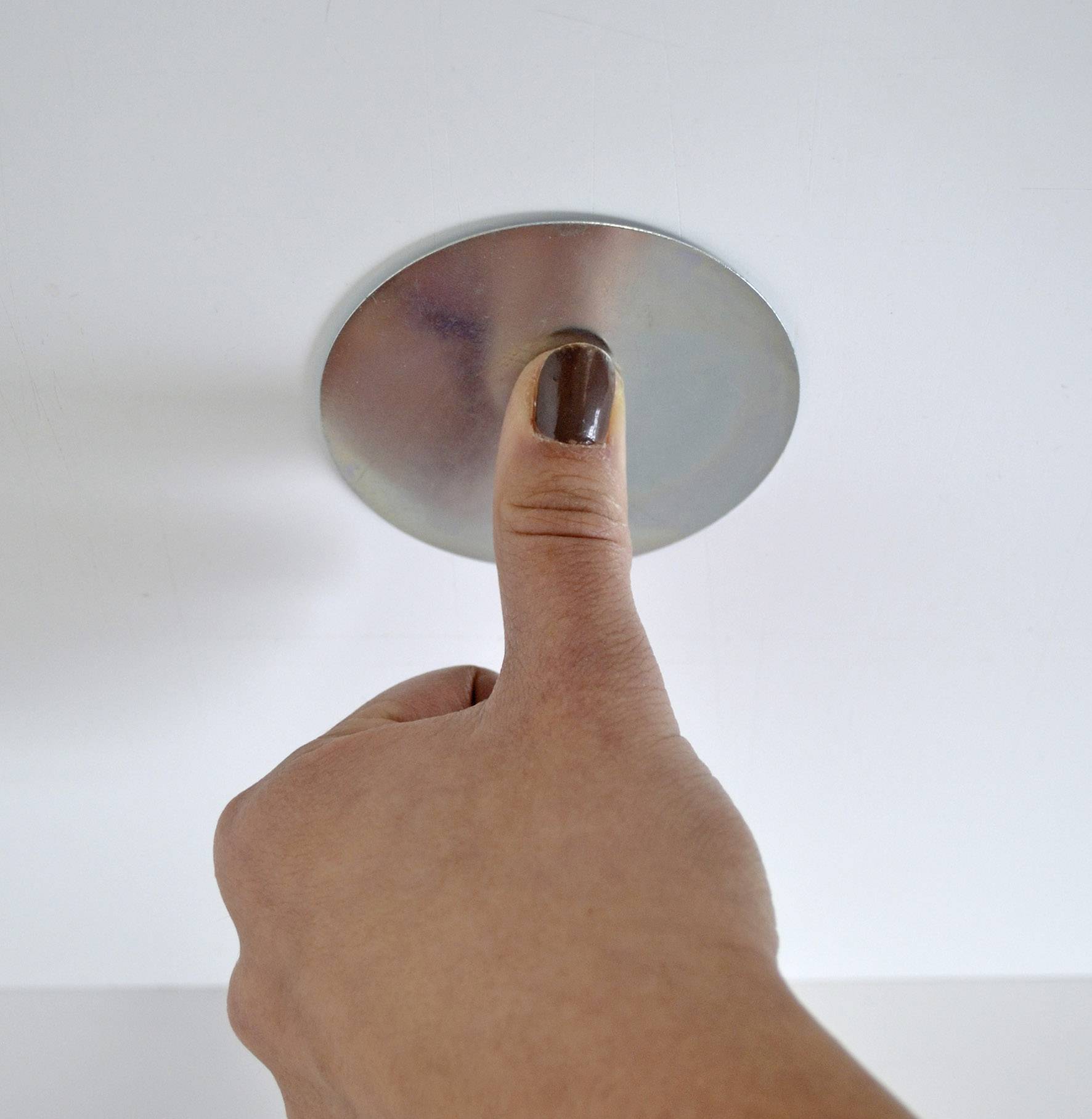Thumb presses against a round, metallic surface on a white wall.