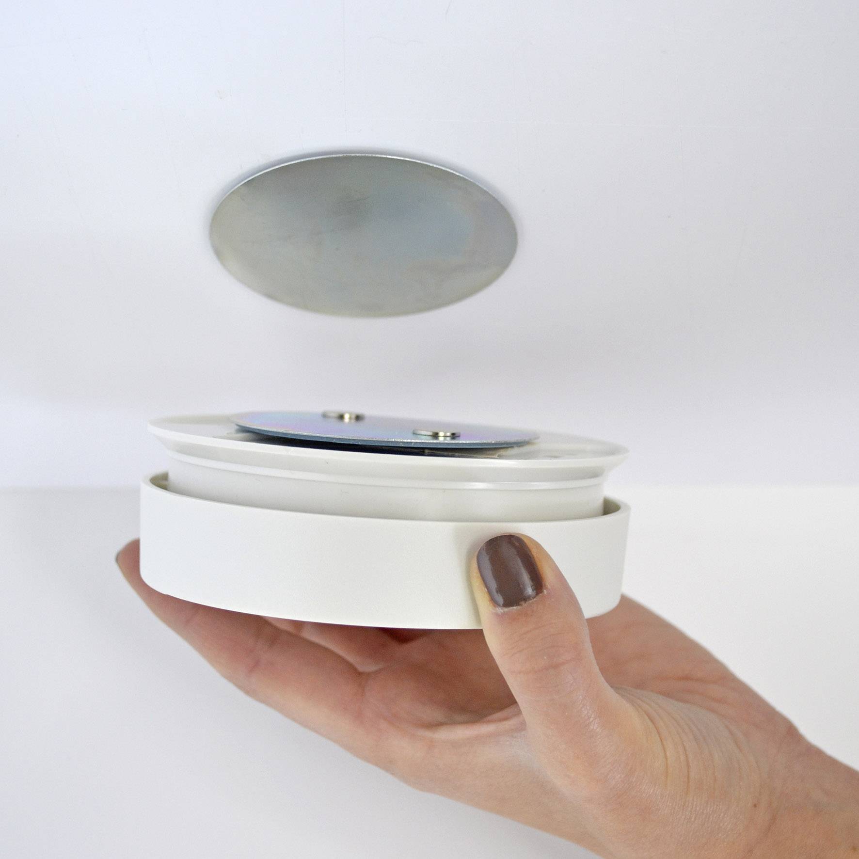 A hand holds a round, white, flat device close to a similar plate mounted on the ceiling. Possibly a magnetic catch.