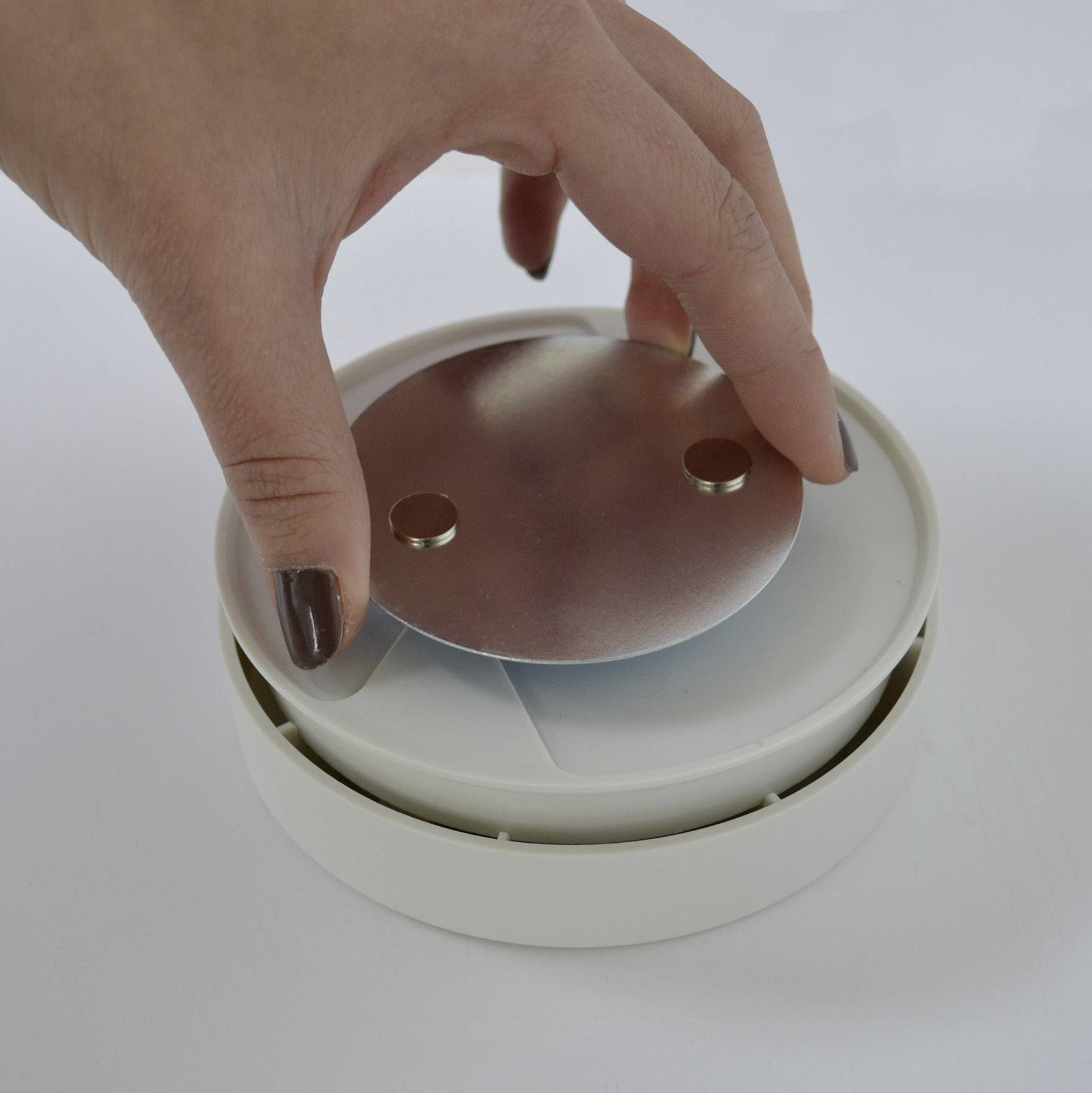A hand is placing a round metal magnet into a white plastic container, possibly a smoke detector or alarm device.