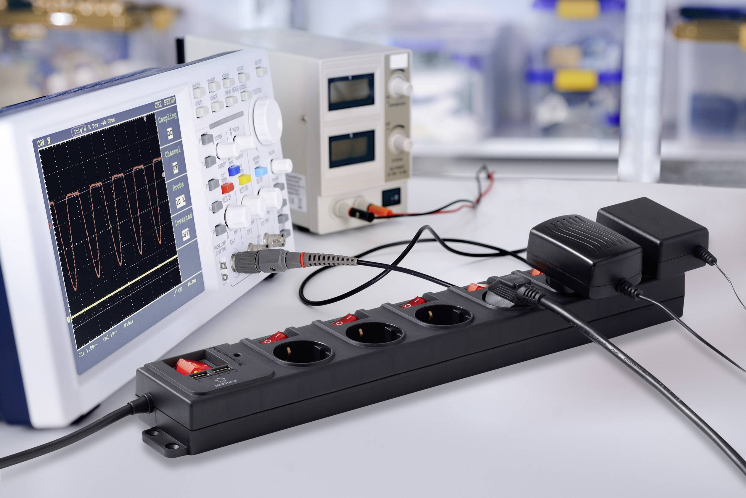 An oscilloscope displays a waveform, alongside a power supply and a multi-socket extension lead with several devices connected.