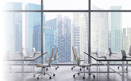 A modern office with empty desks and computers in front of a large window front, overlooking a city skyline.