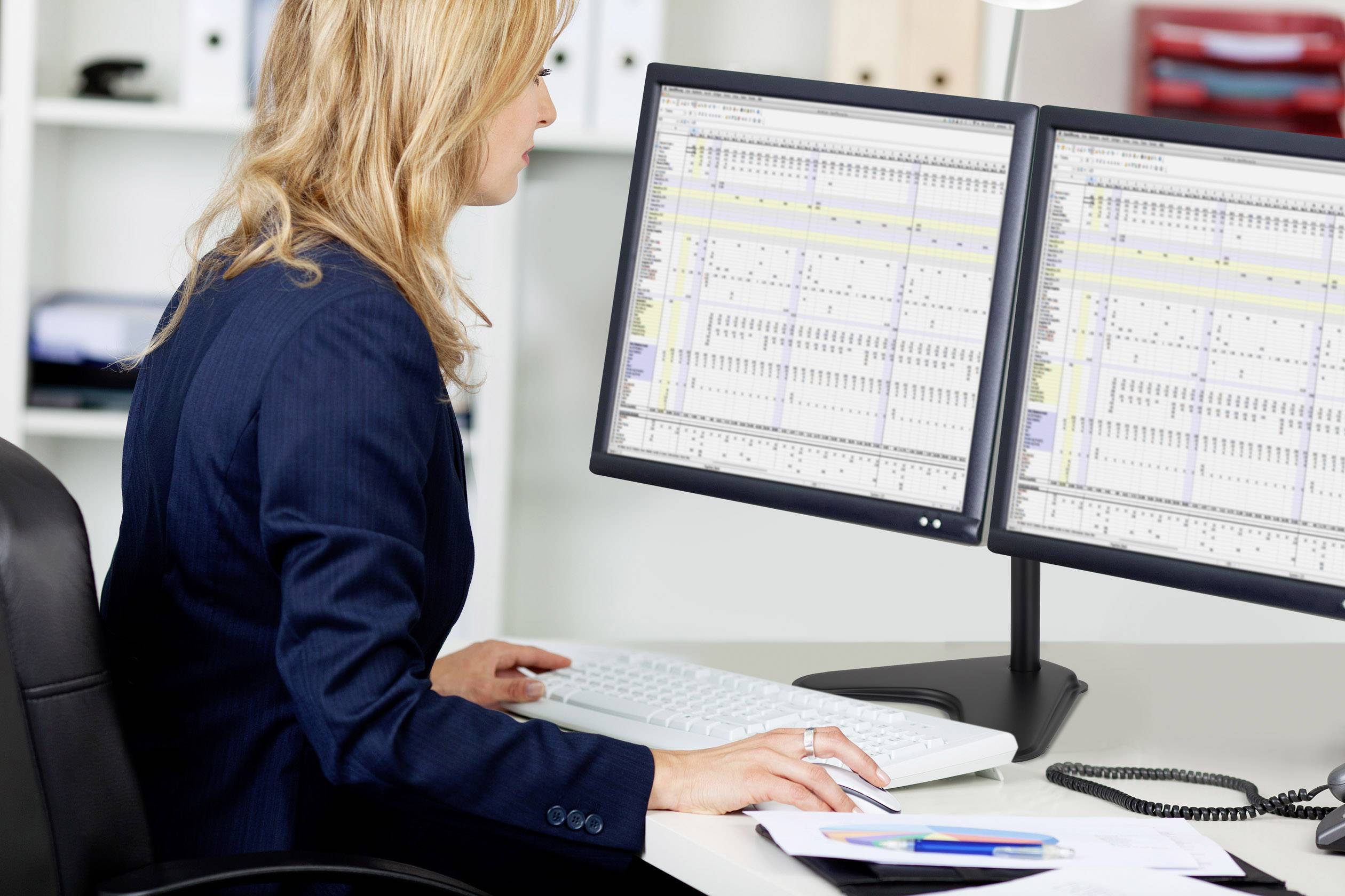 A woman sits at a desk, working on two computer screens with spreadsheets, showing concentration and professional activity.