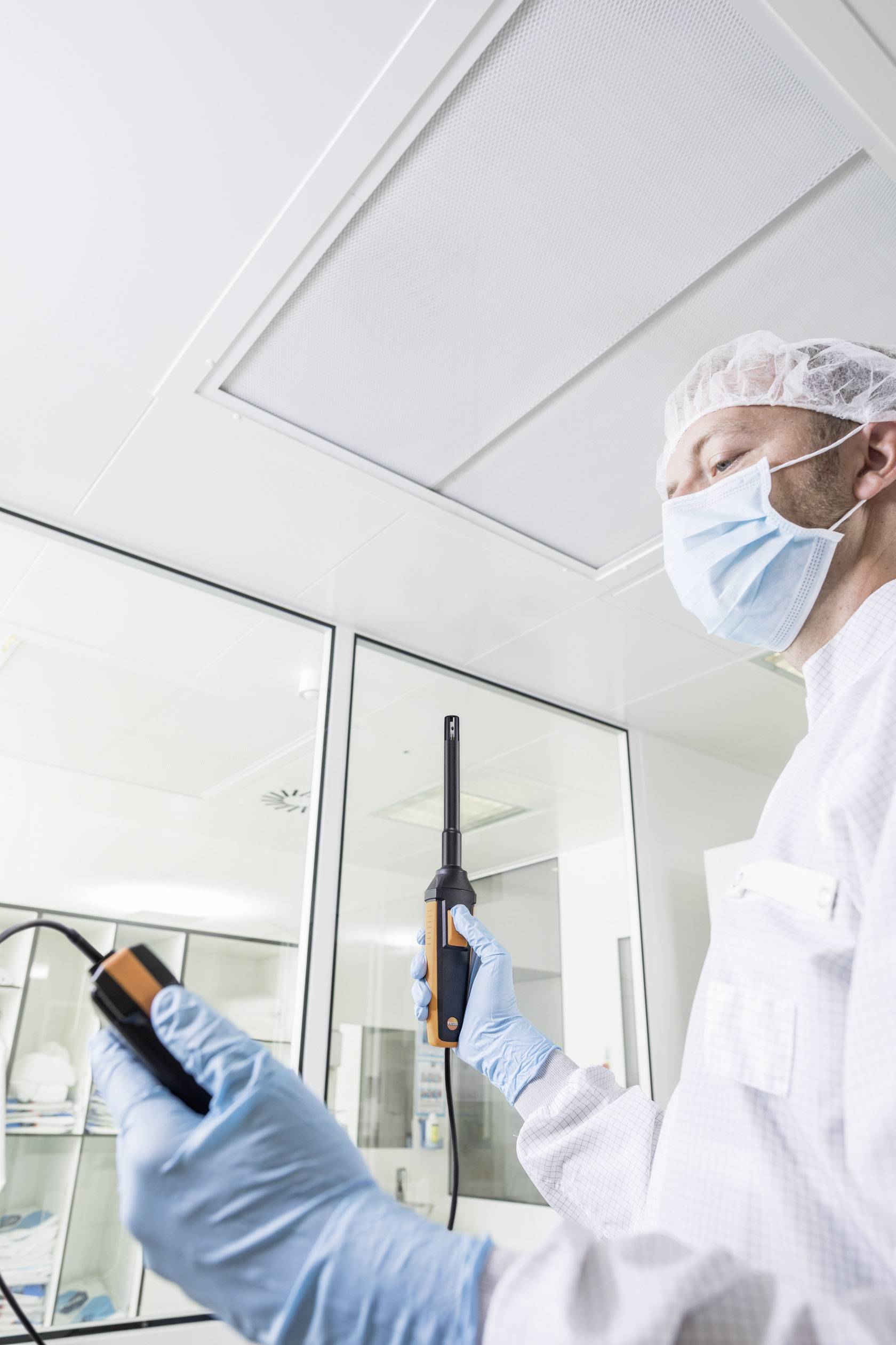 A laboratory technician in sterile clothing measures air quality with a device in a clean room. The background shows a laboratory environment.
