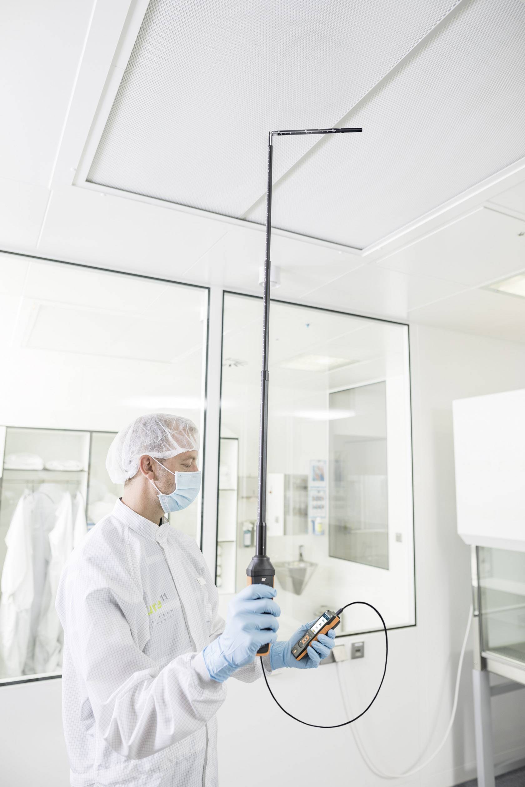 A person in protective clothing is measuring air quality with an electronic device in a clean room.