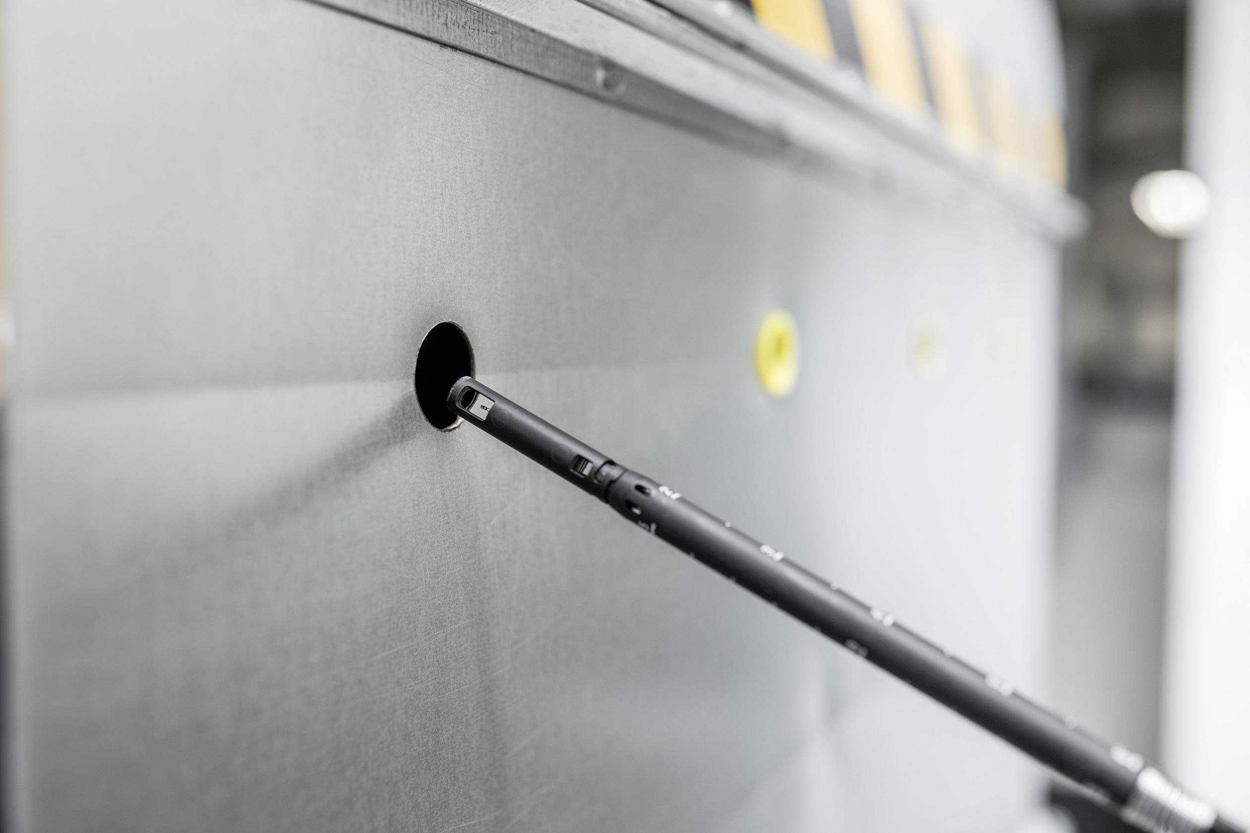 A black metal rod is inserted into a hole in a grey metal surface. Yellow dots are visible underneath.