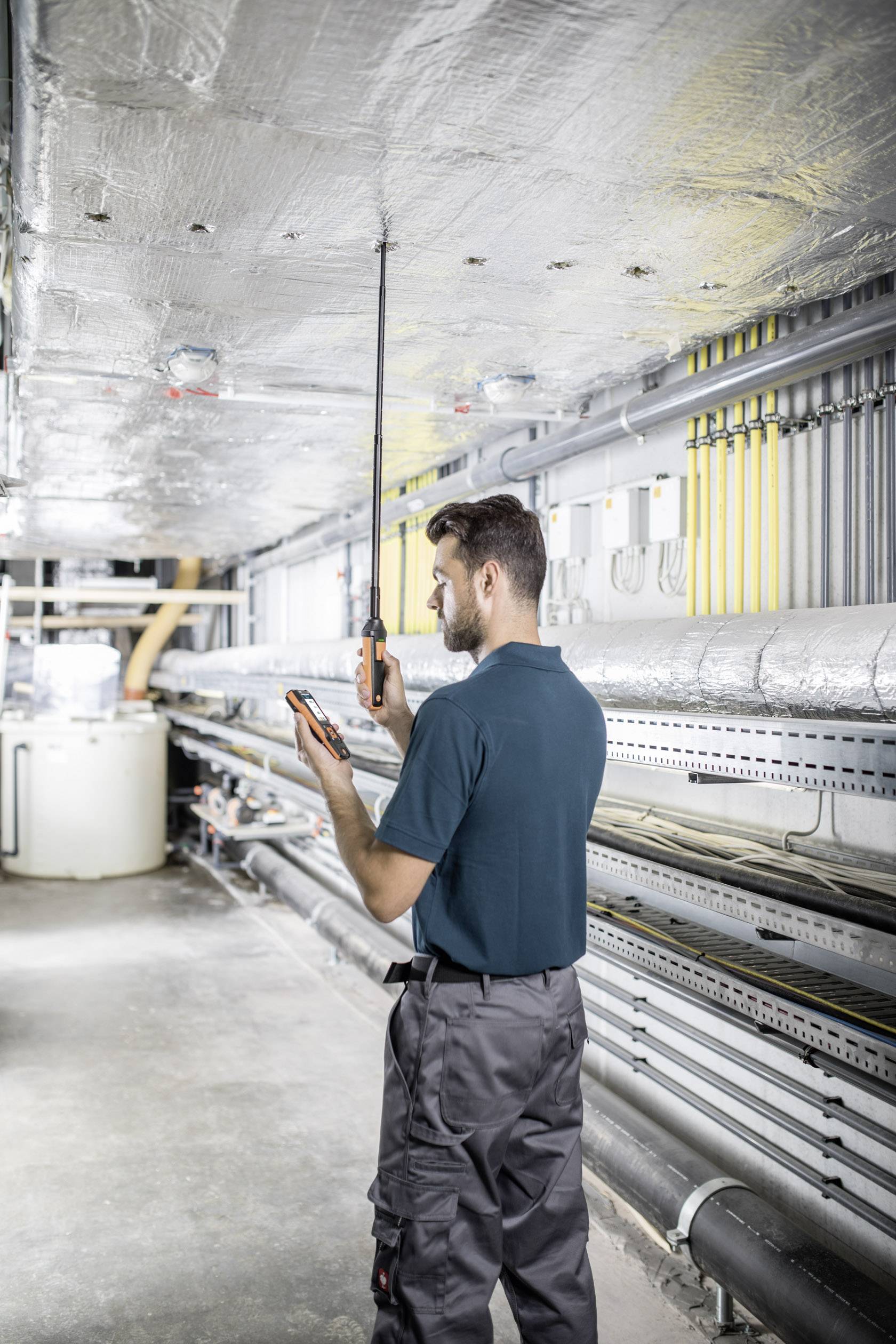 A technician checks the ceiling with a device in an industrial technical environment. Pipes and cables are visible in the background.