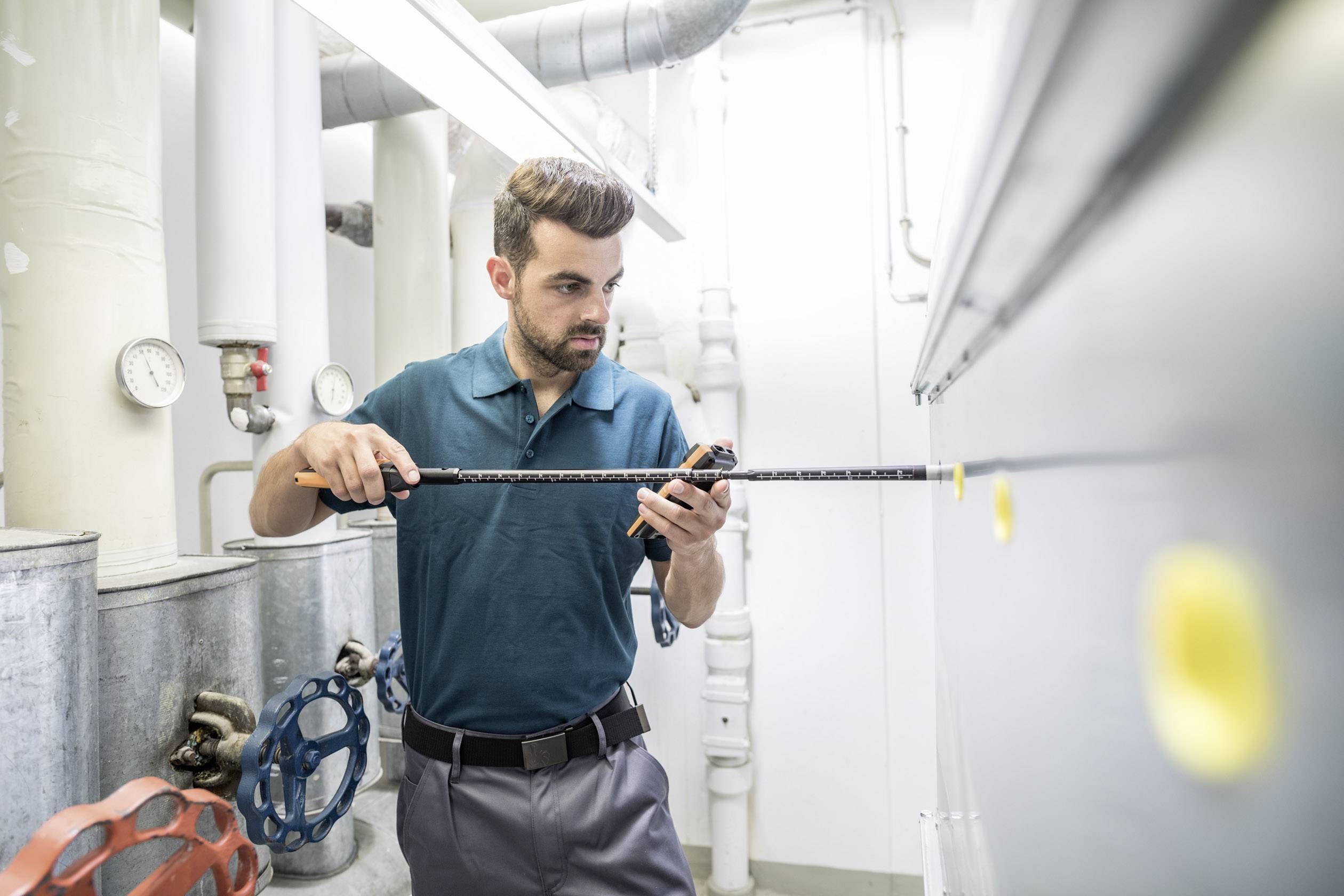 A man is measuring with a tape measure in a technical room with pipes and valves. He is wearing a green shirt and is focused on his work.