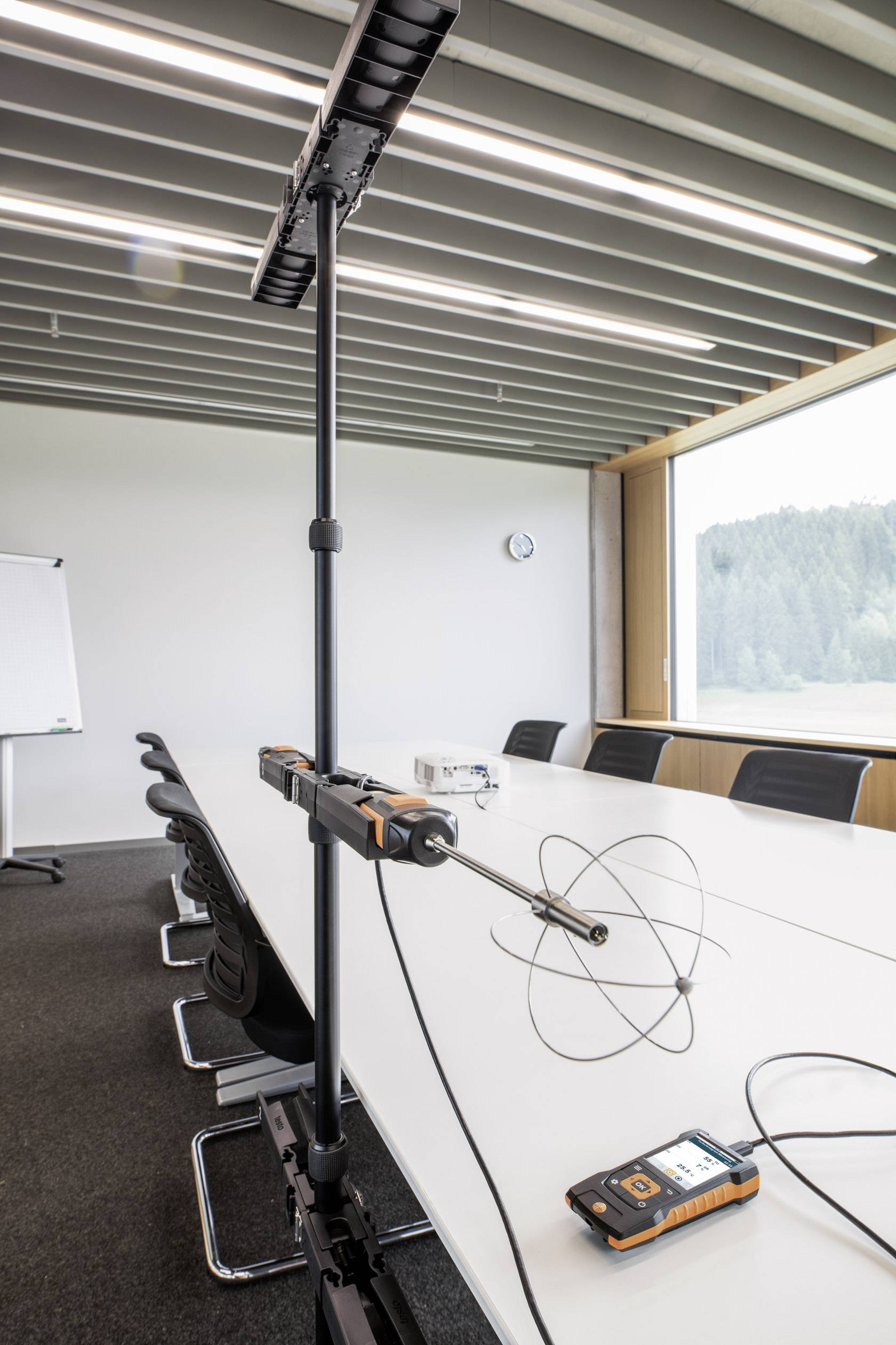 A meeting room with a long table, chairs and a window view. On the table sits a measuring device with an antenna for room acoustics.