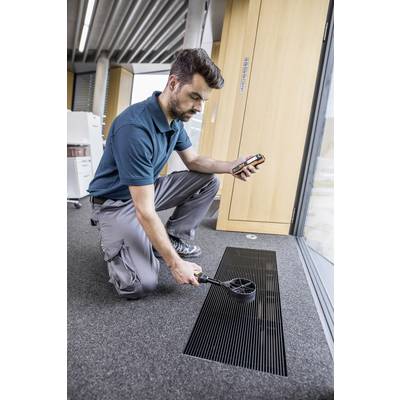 A man is kneeling on a carpet and measuring air quality with a handheld device in front of a ventilation grille in an office.