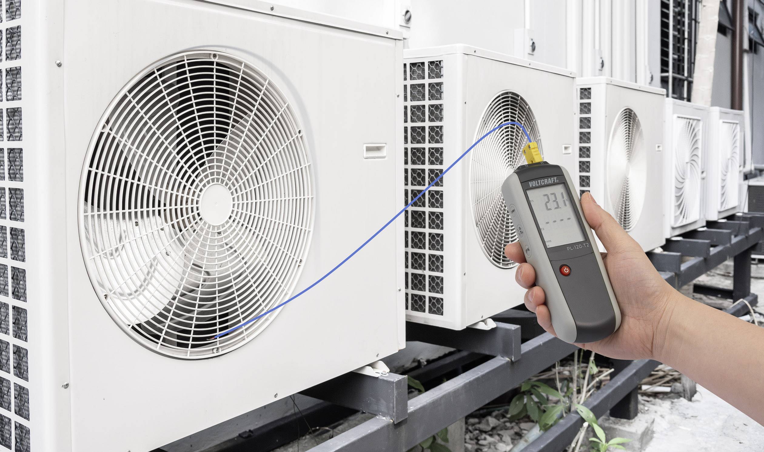 A hand holds a digital thermometer in front of air conditioning units. The display shows '23°C'. The image demonstrates temperature measurement.
