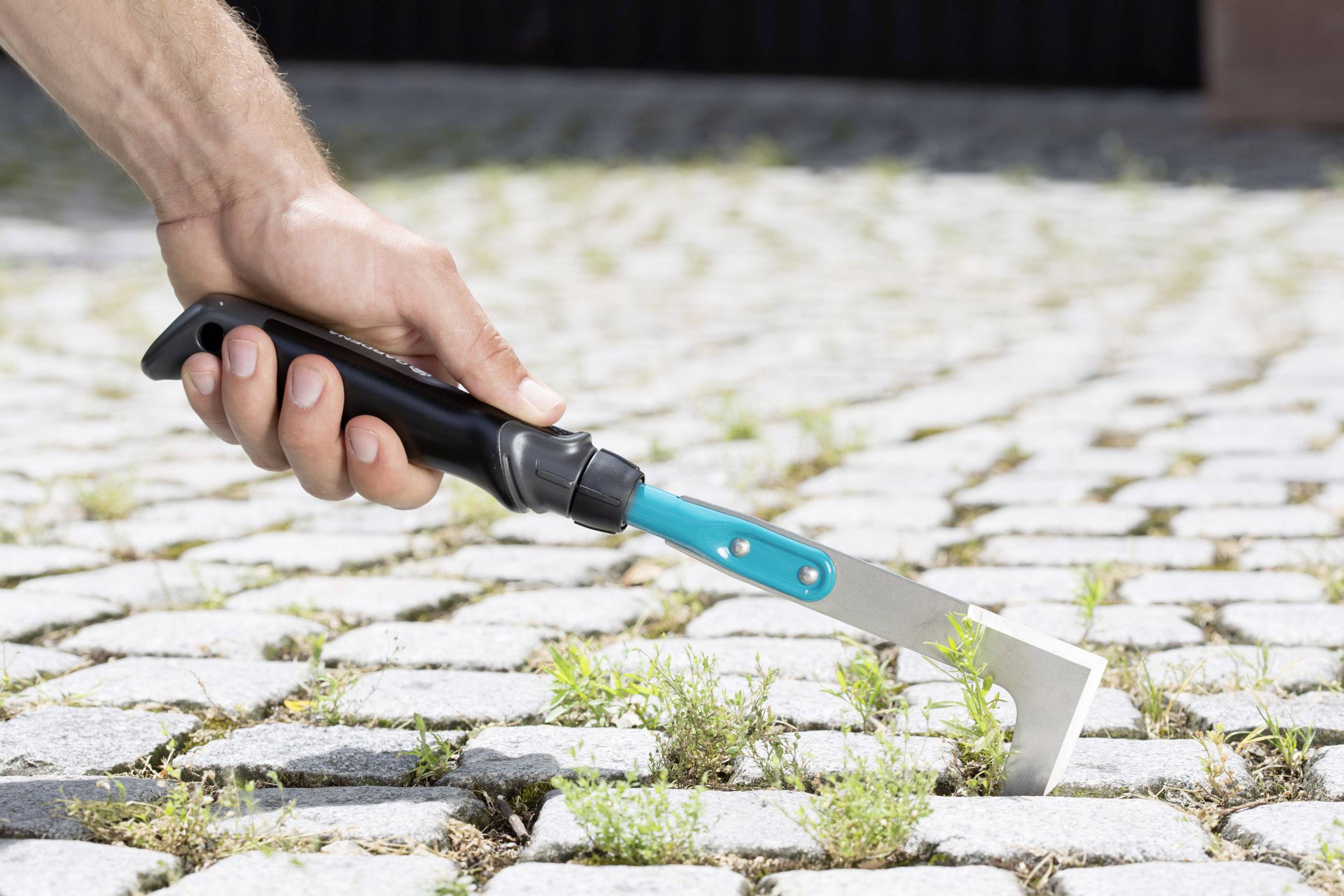 A hand holds a joint scraper tool over a paved pathway covered in weeds, ready to clean the joints.