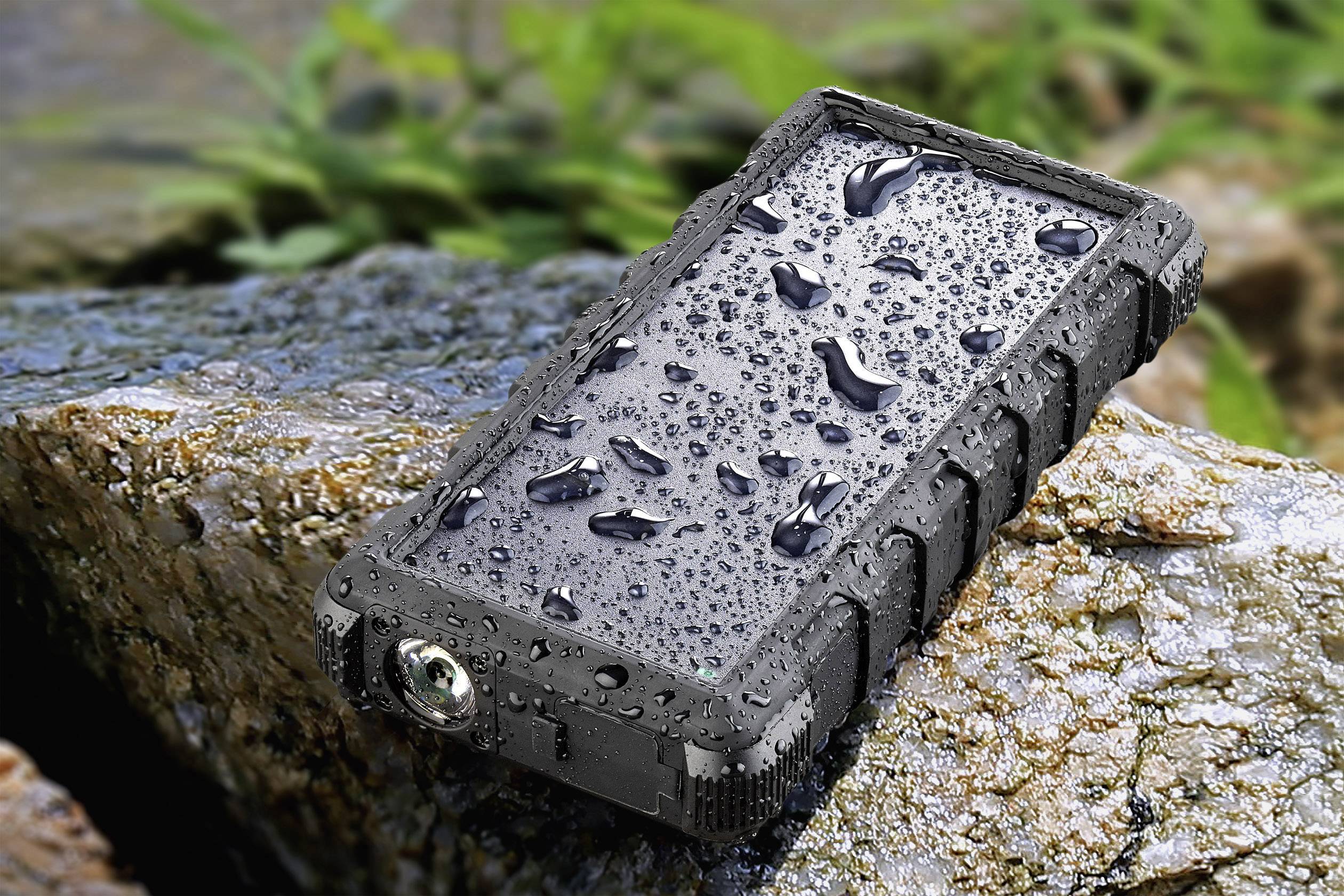 Robust, waterproof device with droplets on its surface lies on a stone, surrounded by leaves.