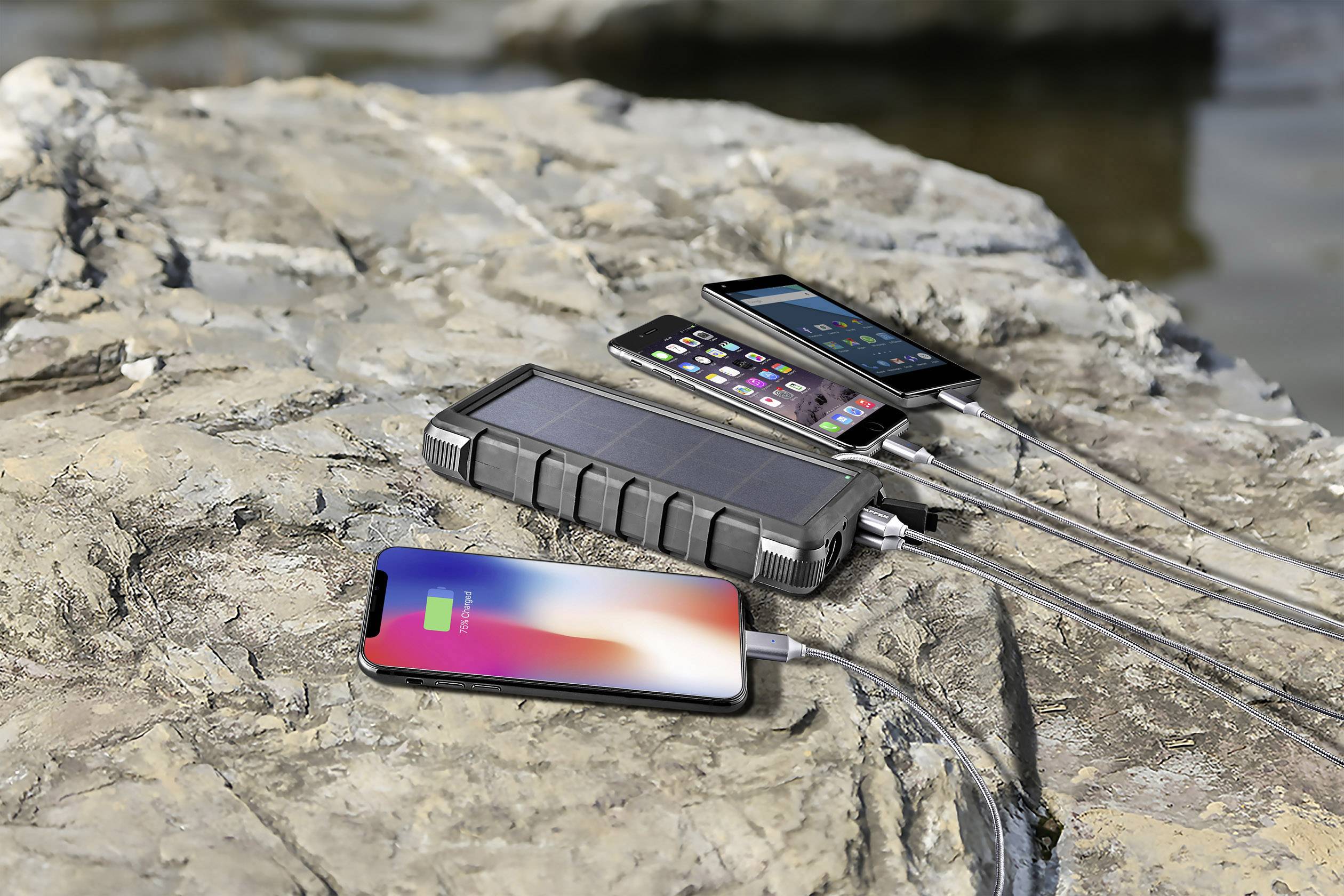 Three smartphones are being charged on a rock using a power bank with a solar panel.