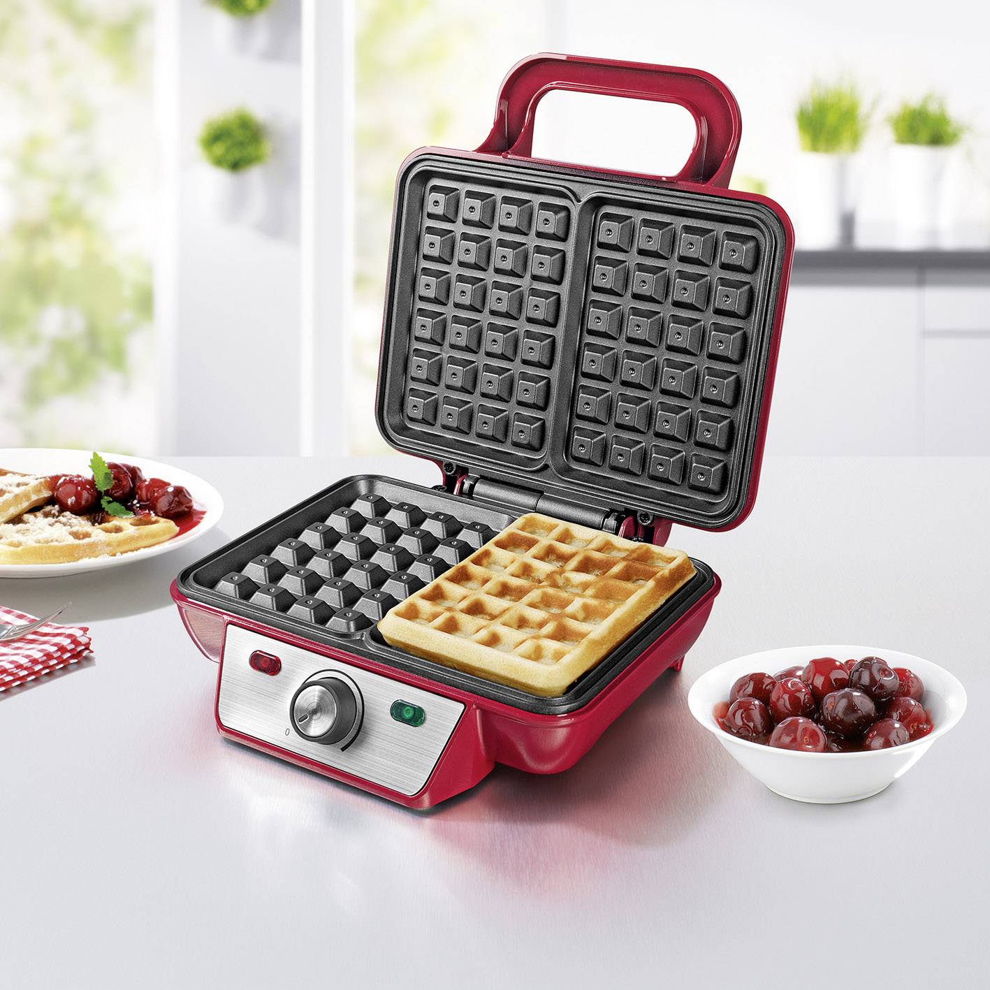 A red waffle maker on a table, with open plates, shows a finished waffle. Beside it are waffles with berries and cream.