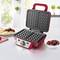 Red waffle iron with open lid on a kitchen worktop, next to fresh waffles and a bowl of cherries.