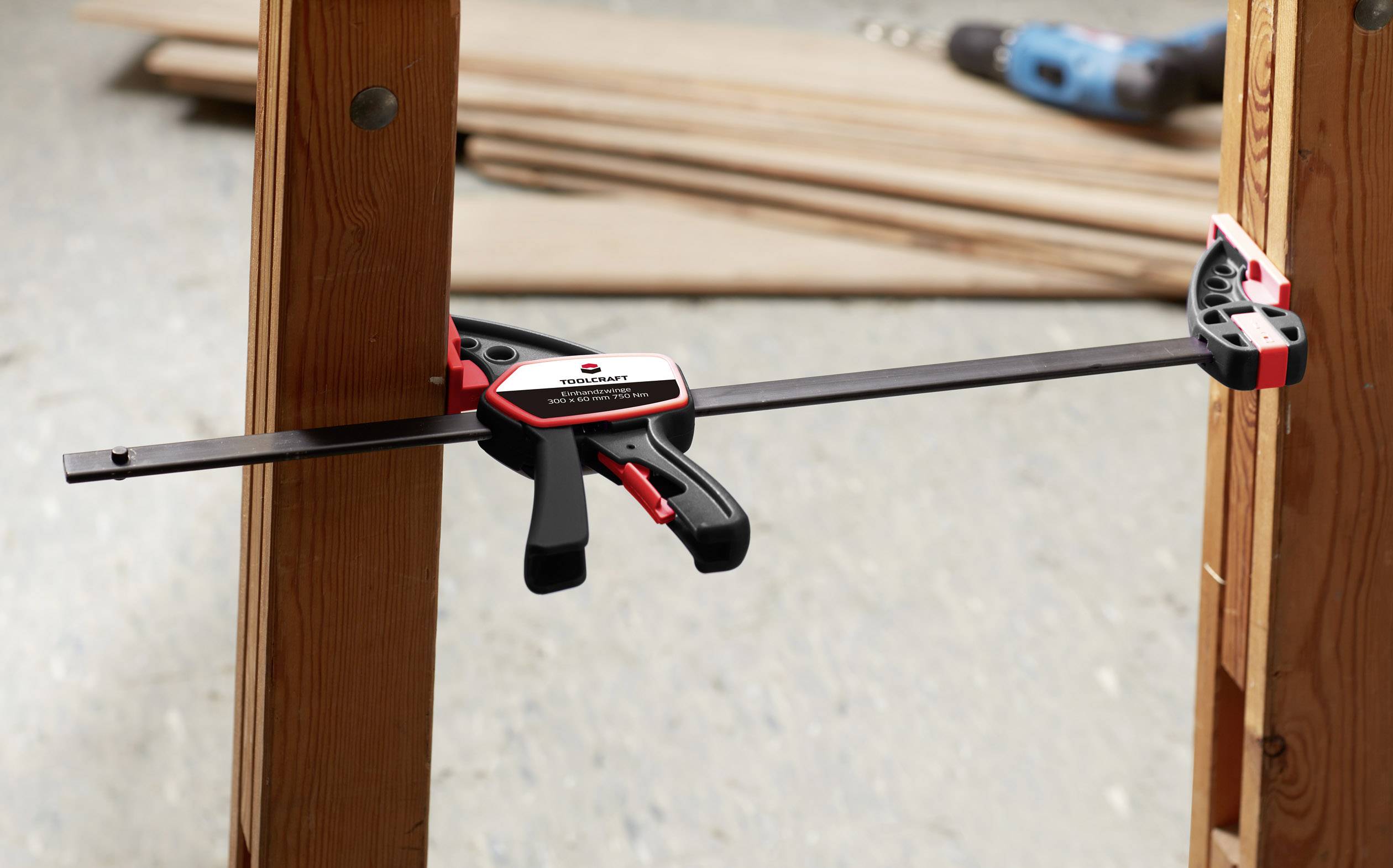 A G-clamp holds two pieces of wood together. In the background, wooden planks and a cordless drill can be seen.