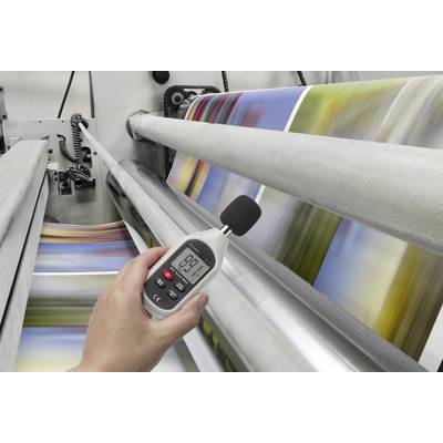 A person is measuring the noise level of a printing press using a decibel meter while it is printing colourful pages.